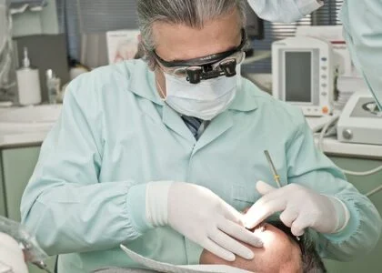 grey heaired dentist with white gloves and green scrubs working on male patient in chair